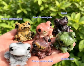 May include: Six small, carved stone frog figurines in different colours and textures. The frogs are labelled with names such as "Chinese painting stone", "Peach blossom jade", and "Garnet". The background is blurred green foliage.