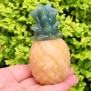 以下が含まれることがあります： 緑色のトップと黄色のボディを持つ、彫刻されたパイナップルの置物。パイナップルは手に置かれています。