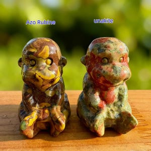 May include: Two small, carved stone monkey figurines. The left one is labeled "Azo Rubine" and features a mix of brown, yellow, and green colors. The right figurine, labeled "unakite," has red, green, and pink hues.