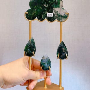 May include: A gold-colored metal stand shaped like a cloud with three hanging raindrops, all made of green moss agate. The cloud piece features a small white sticker with the text D22. The stand has a square base and two vertical support rods.