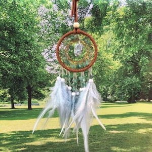 May include: A green and white dream catcher with feathers hanging from the bottom. The dream catcher has a crystal in the center and is decorated with green stones.