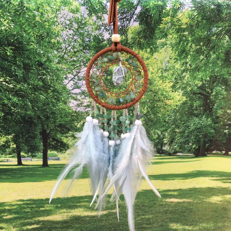 May include: A green and white dream catcher with feathers hanging from the bottom. The dream catcher has a crystal in the center and is decorated with green stones.
