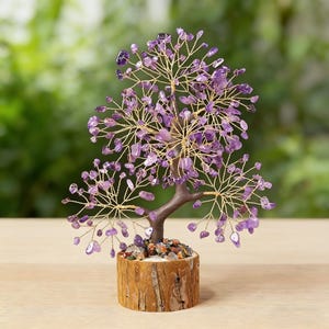 May include: A decorative tree sculpture with a brown trunk and gold-colored wire branches. The branches are adorned with small, purple amethyst stones. The tree sits in a wooden base filled with small, colorful stones. The background is a blurred green.