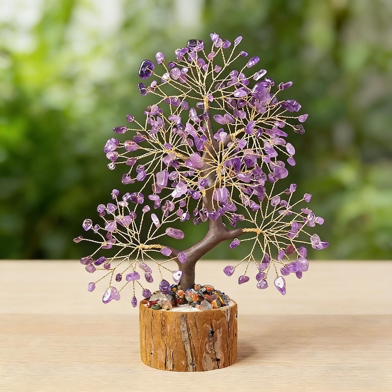 May include: A decorative tree sculpture with a brown trunk and gold-colored wire branches. The branches are adorned with small, purple amethyst stones. The tree sits in a wooden base filled with small, colorful stones. The background is a blurred green.