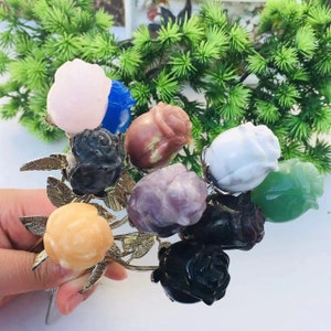 以下が含まれることがあります： ピンク、青、緑など、さまざまな色の彫刻された石のバラのブーケ。 バラは、葉が付いた銀色の金属製の茎に取り付けられています。 このアレンジメントは、緑の葉の背景に対して配置されています。