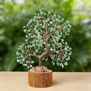 May include: A decorative tree sculpture with a brown trunk and branches, adorned with green gemstone leaves. The tree sits in a wooden base filled with colorful stones. The tree is approximately 6 inches tall.