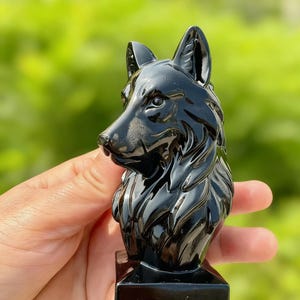 May include: A polished black wolf sculpture, featuring intricate detailing of fur and facial characteristics. The figurine is mounted on a rectangular base. The wolf's head is turned to the side, displaying its pointed ears and attentive gaze. The background is a blurred green.