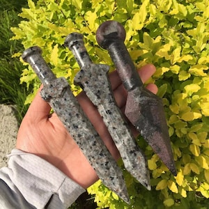 May include: Three decorative swords made of polished gray and black stone. The swords have a pointed tip and a slightly curved blade. The swords are held in a hand.