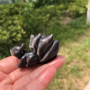 May include: A small, polished obsidian fox figurine. The fox is depicted in a curled position with a detailed tail and body. The figurine has a smooth, reflective surface and is a dark, metallic gray color.