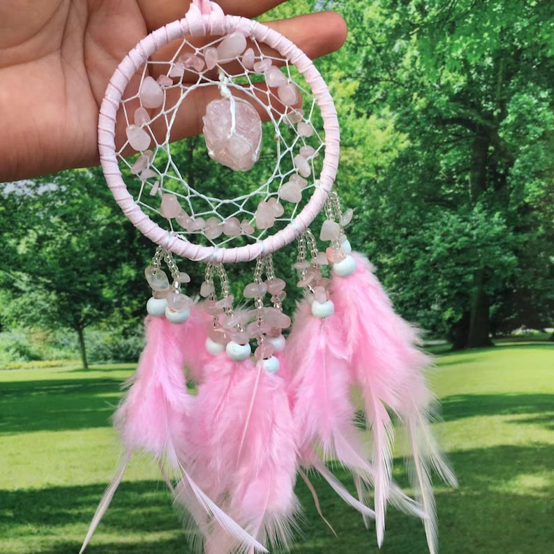 May include: Pink dream catcher with a large rose quartz crystal in the center, surrounded by smaller rose quartz stones. The dream catcher has pink feathers and white beads.
