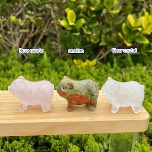 May include: Three carved pig figurines made of rose quartz, unakite, and clear crystal are displayed on a wooden surface. The rose quartz pig is pink, the unakite pig is green and red, and the clear crystal pig is transparent. Text above each pig identifies the material.