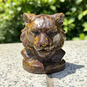 Könnte beinhalten: Eine detaillierte Skulptur eines Tigerkopfes aus Stein mit einem marmorierten Muster in Braun-, Gold- und Rottönen. Die Gesichtszüge des Tigers sind deutlich erkennbar. Die Skulptur steht auf einem runden Sockel.