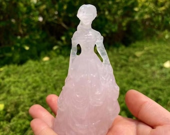 May include: A translucent pale pink rose quartz carving shaped like a standing figure wearing a long, flowing gown. The sculpture features a defined head, torso, and a wide, flared skirt with subtle carved details, showcasing the natural stone texture.