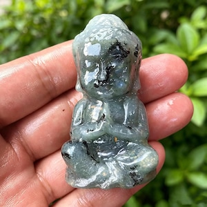May include: A small, seated Buddha statue carved from a translucent, pale green stone. The statue has a serene expression and is in a meditative pose with hands clasped. Darker inclusions are visible within the stone.
