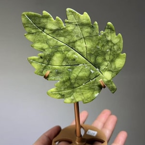 May include: A carved green stone leaf sculpture with dark veining and a serrated edge, mounted on a copper-colored metal rod and a circular base with cutouts. The leaf features a central vein and natural mineral inclusions throughout the surface.
