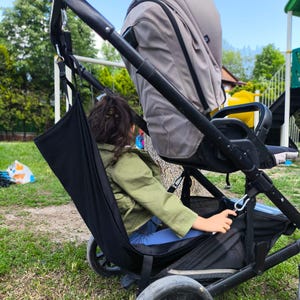 Könnte beinhalten: Eine schwarze Kinderwagen-Aufbewahrungstasche, die am Griff eines Kinderwagens mit beigem Verdeck befestigt ist. Die Tasche besteht aus schwarzem Stoff und hängt vertikal am Kinderwagengestell, was zusätzlichen Stauraum für verschiedene Gegenstände bietet.