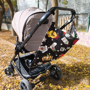 May include: A black stroller organizer bag with a festive Christmas pattern, including snowflakes, gifts, and stockings, hangs from a black stroller. The stroller has a beige canopy and is positioned on a bed of fallen yellow leaves.