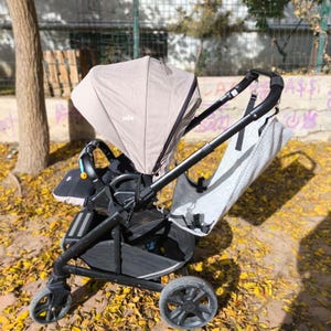 Könnte beinhalten: Ein schwarzer und grauer Kinderwagen mit einem beigen Verdeck und einem gemusterten Sitzbezug. Der Kinderwagen hat vier Räder und einen Stauraum darunter. Der Markenname "Joie" ist auf dem Verdeck sichtbar. Der Kinderwagen steht auf einer Oberfläche mit gelben Blättern.