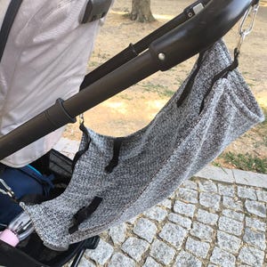 May include: A gray stroller organizer bag with black accents is attached to the stroller handle. The bag is made of a textured fabric and hangs from the stroller with metal clips. A water bottle is visible in the stroller's storage area.
