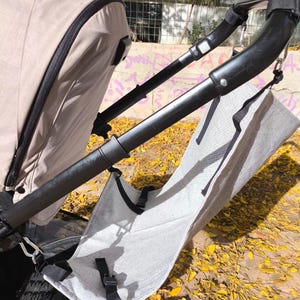 Può includere: Un passeggino beige con manici neri e un accessorio in tessuto grigio. Il passeggino ha una tendina parasole e un'area portaoggetti in rete. L'immagine è scattata all'aperto con foglie gialle sul terreno.