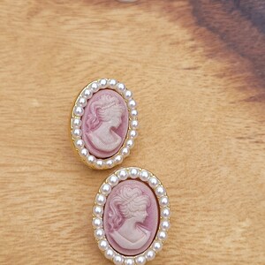 Pu&ograve; includere: Un paio di orecchini ovali con un design a cammeo. I cammei presentano un profilo rosa e bianco di un viso femminile. Gli orecchini sono circondati da un bordo di perle bianche.