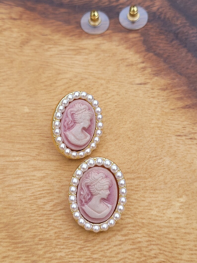 Pu&ograve; includere: Un paio di orecchini ovali con un design a cammeo. I cammei presentano un profilo rosa e bianco di un viso femminile. Gli orecchini sono circondati da un bordo di perle bianche.