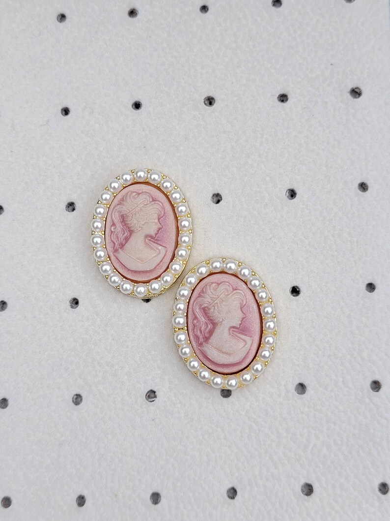 Pu&ograve; includere: Un paio di orecchini con cammeo rosa con bordo dorato e bordo di perle.