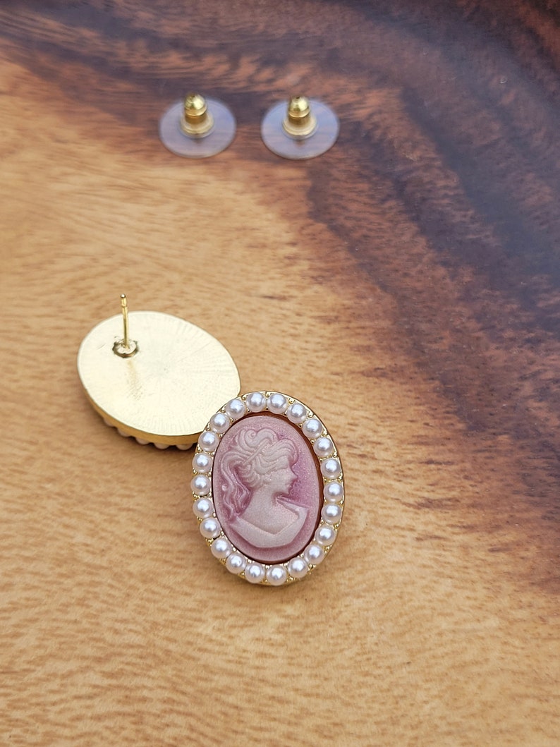 Pu&ograve; includere: Un paio di orecchini dorati con un design a cammeo. Il cammeo presenta il profilo di una donna in rosa e bianco. Gli orecchini sono circondati da un cerchio di perle bianche.
