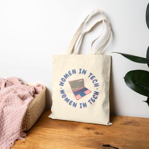 May include: A cream-colored tote bag with the text "WOMEN IN TECH" encircling a pink and blue laptop graphic. The bag has two long handles and is made of a sturdy fabric. The design is centered on the front of the bag.