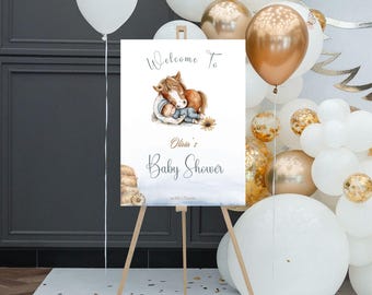 May include: Welcome to Olivia's Baby Shower sign with a watercolor illustration of a brown horse and a baby sleeping on its back. The sign is on a wooden easel in front of a white and gold balloon backdrop.