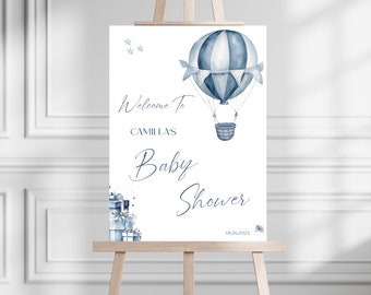 May include: A white welcome sign with blue text and illustrations for a baby shower. The sign says "Welcome to Camilla's Baby Shower" with a date of 08.26.2023. The illustration is of a blue and white hot air balloon with a basket and stars.