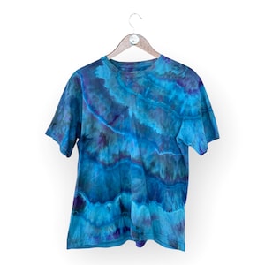 Pode incluir: Uma camiseta azul tie-dye com um padrão de redemoinho. A camiseta tem mangas curtas e gola redonda.