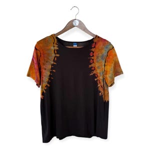 Op de afbeelding: Zwart T-shirt met korte mouwen en tie-dye accenten in tinten oranje, geel en roze. Het shirt hangt aan een houten hanger tegen een witte achtergrond. Het tie-dye patroon bevindt zich op de schouders en mouwen.