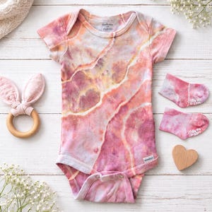 May include: A tie-dye baby bodysuit and socks set in shades of pink, peach, and yellow. The short-sleeved bodysuit has a snap closure. A wooden bunny ear teether and a wooden heart are also in the image.