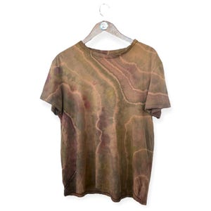 Pode incluir: Uma camiseta tie-dye marrom, verde e vermelha com um padrão de redemoinho. A camiseta está pendurada em um cabide de madeira.