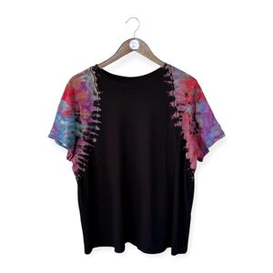 May include: Black t-shirt with tie-dye sleeves in shades of pink, purple, and blue. The short-sleeved shirt is hanging on a wooden hanger against a white background. The tie-dye pattern is vertical along the sleeves.