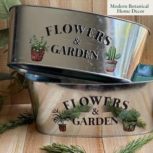 May include: Two galvanized metal planters with an oval shape. The planters have the words "FLOWERS & GARDEN" printed on them in black, along with illustrations of potted plants. The planters are for home decor.