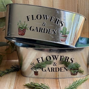 May include: Two galvanized metal oval planters. Each planter has the words "FLOWERS & GARDEN" printed in black. The planters are decorated with images of potted plants, including a cactus and other greenery.