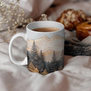 May include: White ceramic mug with a mountain and forest scene. The mountains are depicted in shades of beige and gold, with a forest of dark green trees in the foreground.
