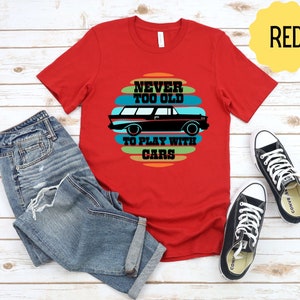 Puede incluir: Camiseta roja con un gr&aacute;fico de coche vintage y el texto "Never Too Old To Play With Cars".