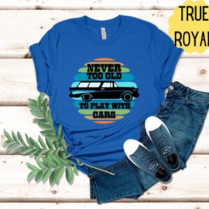 Puede incluir: Una camiseta azul real con un gr&aacute;fico de coche vintage y el texto "Never too old to play with cars". El texto est&aacute; en una fuente retro y est&aacute; rodeado de rayas de colores.