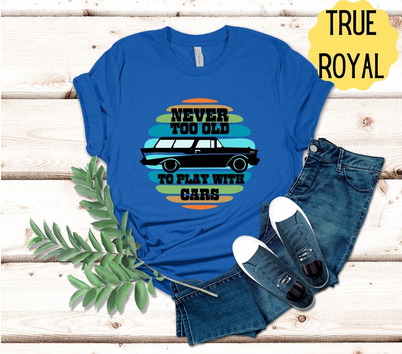 Puede incluir: Una camiseta azul real con un gr&aacute;fico de coche vintage y el texto "Never too old to play with cars". El texto est&aacute; en una fuente retro y est&aacute; rodeado de rayas de colores.
