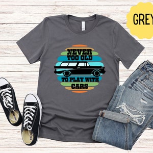 Puede incluir: Una camiseta gris con un gr&aacute;fico de coche vintage y el texto "Never Too Old To Play With Cars".