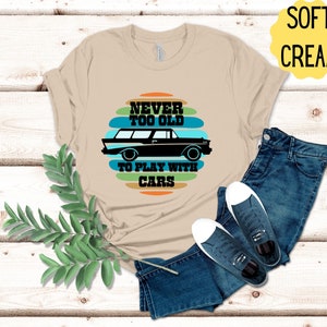 Puede incluir: Una camiseta beige con un gr&aacute;fico de coche vintage y el texto "Never too old to play with cars" en un dise&ntilde;o a rayas de colores.