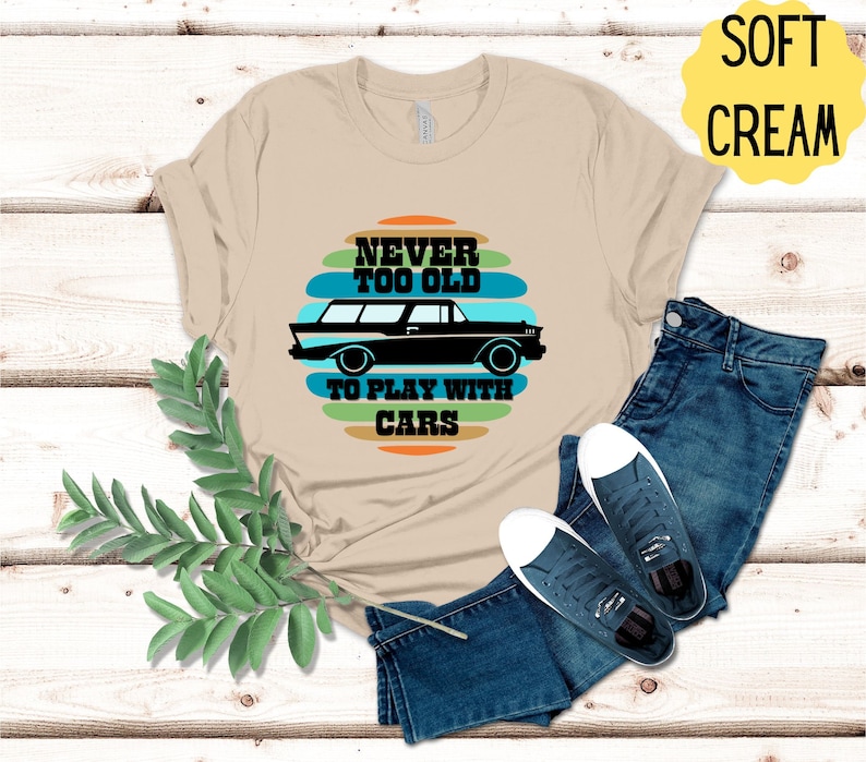 Puede incluir: Una camiseta beige con un gr&aacute;fico de coche vintage y el texto "Never too old to play with cars" en un dise&ntilde;o a rayas de colores.