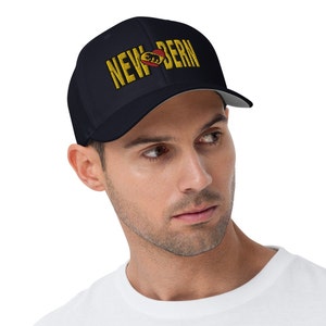 Könnte beinhalten: Eine marineblaue Baseballkappe mit weißem Schirm. Auf der Vorderseite der Kappe befindet sich ein goldfarben gesticktes Design mit der Aufschrift "NEW BERN" mit einem roten Herzen und einem Bären im Herzen.