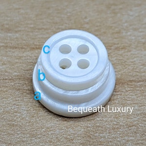 May include: White button with four holes, a raised rim, and a textured base. The button is labeled "Bequeath Luxury".