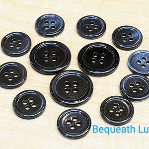 May include: A collection of 12 black plastic buttons with four holes each. The buttons are arranged in a circle on a light brown surface. The text "Bequeath Luxury" is visible in the bottom right corner of the image.