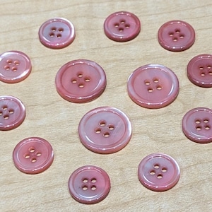 May include: A collection of pink buttons with four holes each, arranged on a light brown wooden surface. The buttons vary in size, with a pearlescent sheen. Ideal for sewing and craft projects.