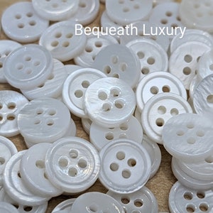 May include: A close-up of a pile of white, four-hole, mother-of-pearl buttons. The buttons are smooth and shiny, with a slight iridescent sheen.